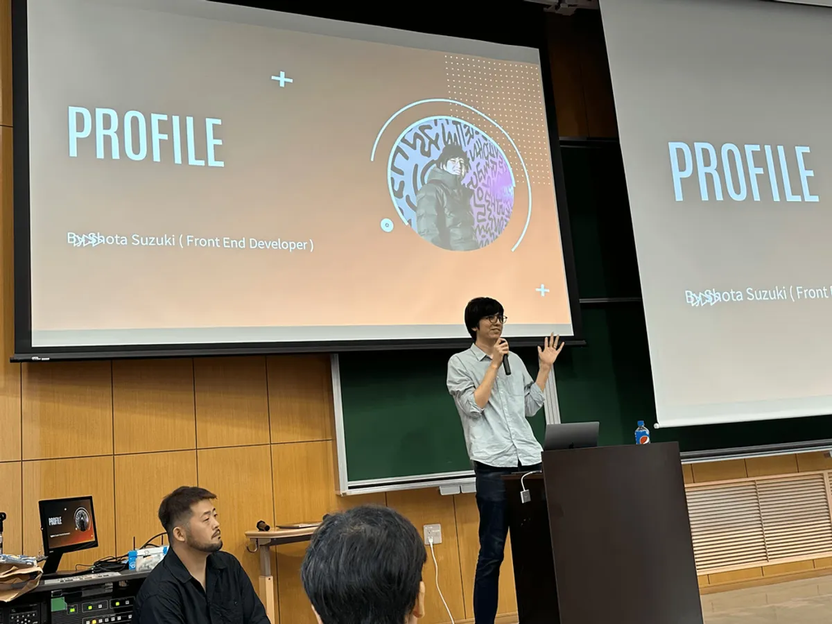 A D&S front-end developer speaking into a microphone in a Saga University lecture hall, his profile slide shown on the screens behind him