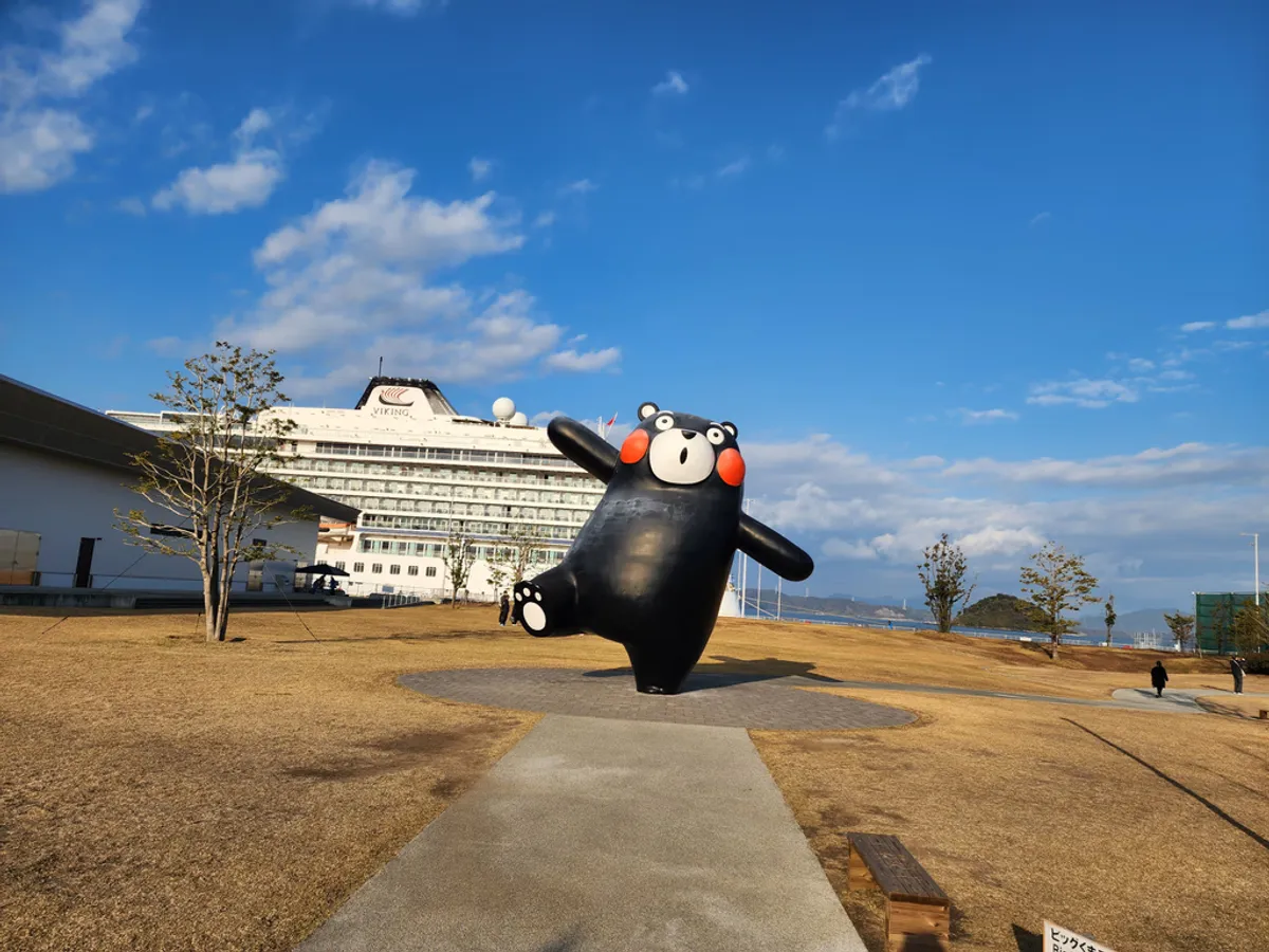 A large Kumamon statue on a waterfront lawn with a cruise ship docked behind it under a clear blue sky