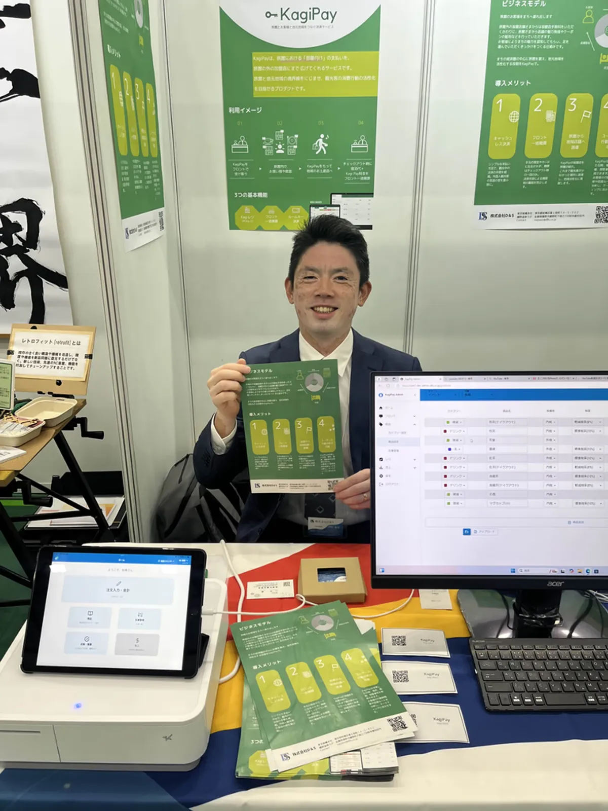 A D&S representative at the KagiPay booth holding a product leaflet, with a tablet and the KagiPay admin screen on the table